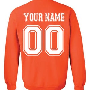 May include: An orange sweatshirt with the text "YOUR NAME" in white block letters above the number "00". The numbers are outlined in white. The sweatshirt has long sleeves and a crew neck.