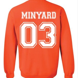 May include: Orange sweatshirt with the name "MINYARD" in white block letters across the upper back. Below the name is the number "03" in large white block letters. The sweatshirt has long sleeves and a crew neck.