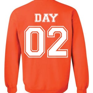 May include: Orange sweatshirt with white text that reads "DAY 02" on the back. The text is in a collegiate font. The sweatshirt has long sleeves and a crew neck.