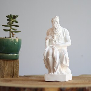 Peut inclure: Statue en plâtre blanc d'une figure assise avec une longue barbe et un livre sur ses genoux. La statue est sur un socle en bois et une petite plante en pot est au premier plan.
