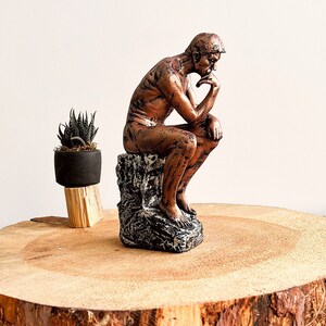 K&ouml;nnte beinhalten: Eine bronzefarbene Skulptur des Denkers, sitzend auf einem strukturierten Sockel, mit einer kleinen Topfpflanze. Die Skulptur steht auf einer gro&szlig;en, runden Holzplatte mit sichtbarer Maserung.