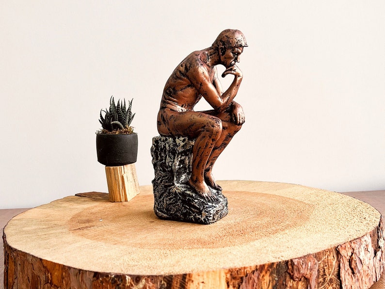 K&ouml;nnte beinhalten: Eine bronzefarbene Skulptur des Denkers, sitzend auf einem strukturierten Sockel, mit einer kleinen Topfpflanze. Die Skulptur steht auf einer gro&szlig;en, runden Holzplatte mit sichtbarer Maserung.