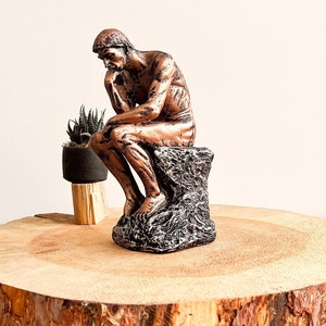 K&ouml;nnte beinhalten: Eine bronzefarbene Skulptur einer sitzenden Figur, der Denker, mit dem Kinn auf der Hand. Die Statue steht auf einem strukturierten, dunkelgrauen Sockel. Eine kleine Topfpflanze steht hinter der Statue auf einer Holzoberfl&auml;che.
