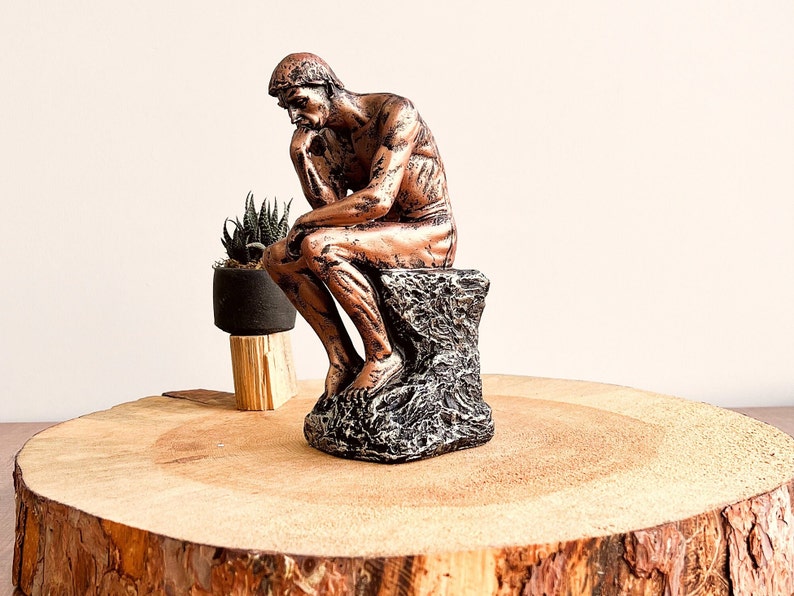 K&ouml;nnte beinhalten: Eine bronzefarbene Skulptur einer sitzenden Figur, der Denker, mit dem Kinn auf der Hand. Die Statue steht auf einem strukturierten, dunkelgrauen Sockel. Eine kleine Topfpflanze steht hinter der Statue auf einer Holzoberfl&auml;che.