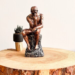K&ouml;nnte beinhalten: Eine bronzefarbene Skulptur des Denkers, sitzend auf einem felsartigen Sockel. Die Statue ist mit einer Hand am Kinn in kontemplativer Haltung dargestellt. Eine kleine Topfpflanze steht links von der Statue auf einem Holzst&auml;nder. Die Skulptur wird auf einer Holzplatte pr&auml;sentiert.