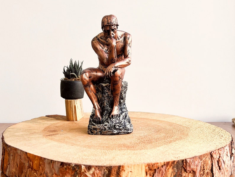 K&ouml;nnte beinhalten: Eine bronzefarbene Skulptur des Denkers, sitzend auf einem felsartigen Sockel. Die Statue ist mit einer Hand am Kinn in kontemplativer Haltung dargestellt. Eine kleine Topfpflanze steht links von der Statue auf einem Holzst&auml;nder. Die Skulptur wird auf einer Holzplatte pr&auml;sentiert.