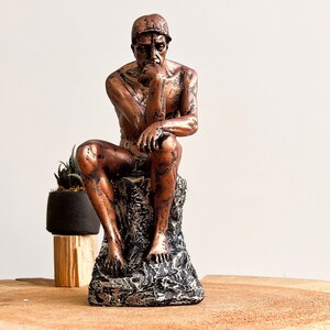 K&ouml;nnte beinhalten: Eine bronzefarbene Skulptur einer sitzenden Figur, die an Rodins "Denker" erinnert. Die Statue steht auf einem strukturierten, felsartigen Sockel. Links von der Skulptur befindet sich eine kleine Topfpflanze.