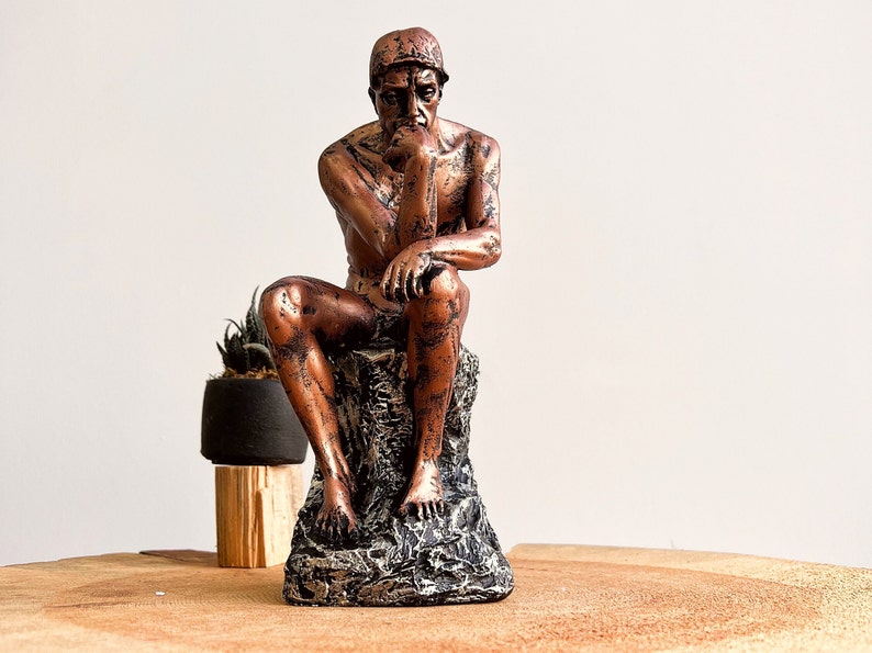 K&ouml;nnte beinhalten: Eine bronzefarbene Skulptur einer sitzenden Figur, die an Rodins "Denker" erinnert. Die Statue steht auf einem strukturierten, felsartigen Sockel. Links von der Skulptur befindet sich eine kleine Topfpflanze.