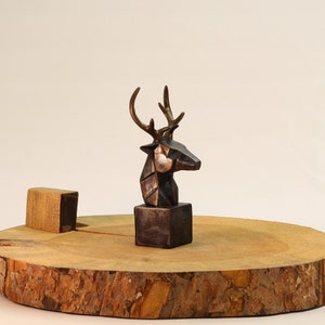 Puede incluir: Una escultura de cabeza de ciervo de color bronce con características geométricas, montada sobre una base cuadrada. La escultura se exhibe sobre una rodaja de madera.