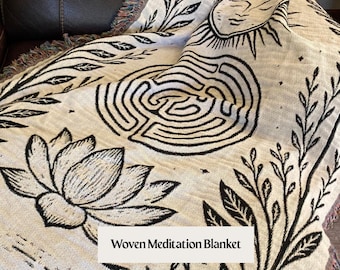 May include: Cream-colored woven blanket with black botanical and celestial designs. Features a lotus flower, sun with a face, and a labyrinth pattern. The text "Woven Meditation Blanket" is displayed on the blanket.