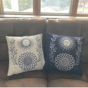 May include: Two decorative throw pillows with celestial designs. One pillow is off-white, the other is navy blue. Each features a sun with a smiling face, a floral border, and a geometric mandala. The pillows are square.