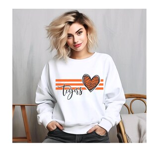 May include: White sweatshirt with orange stripes and a tiger print heart design. The word "Tigers" is written in a script font below the heart. A casual, comfortable top.