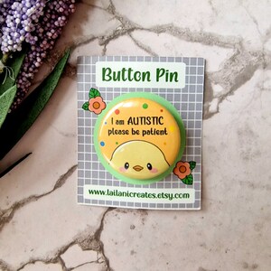 May include: A yellow button pin with a cartoon chick and the text "I am AUTISTIC please be patient". The pin is on a white card with the text "Button Pin" and a website address.