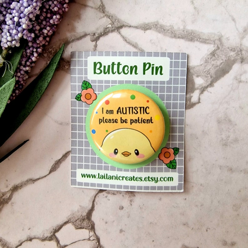 May include: A yellow button pin with a cartoon chick and the text "I am AUTISTIC please be patient". The pin is on a white card with the text "Button Pin" and a website address.