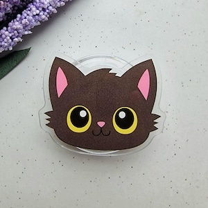 Op de afbeelding: Een doorzichtige, katvormige sticker met een bruin gezicht, roze oren en gele ogen. De sticker heeft een kleine roze neus en een lachende mond. De sticker ligt op een wit oppervlak met paarse bloemen op de achtergrond.