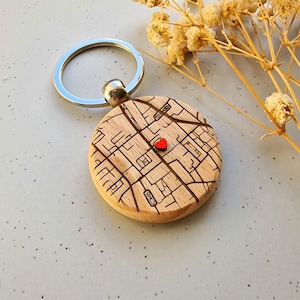 May include: A wooden keyring featuring a circular map design with a small red heart. The keyring has a silver-coloured ring and a silver-coloured attachment. The map is etched into the wood, showing streets and buildings.