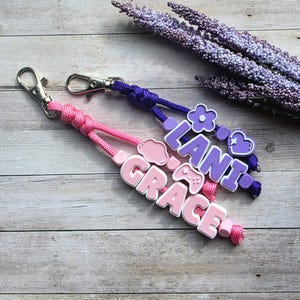 Puede incluir: Dos llaveros personalizados, uno rosa y otro morado. El llavero rosa dice "GRACE" con un encanto de nube y mando de juego. El llavero morado dice "LANI" con un encanto de flor y corazón. Ambos tienen cierres plateados.