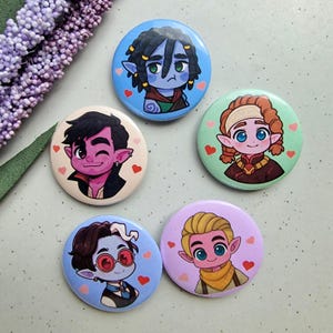 May include: Five round pin badges with cartoon elf characters. Each badge displays a different character design with various skin tones, hair colours, and outfits. The badges are decorated with small heart shapes.