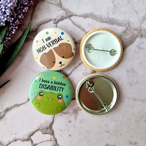 May include: Two button pins with colorful backgrounds and cartoon animals. One pin features a green frog with the text "I have a hidden disability". The other pin features a brown dog with the text "I am non-verbal".
