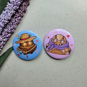 May include: Two round pinback buttons. One button has a blue background with a cartoon character wearing a straw hat. The other button has a purple background with a cartoon character wearing a scarf. Both buttons have heart accents.