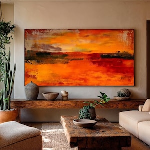 Könnte beinhalten: Ein abstrakter Kunstdruck mit einer lebendigen orange-roten Farbpalette dominiert die Wand. Das Kunstwerk befindet sich über einer Holzkonsolentisch mit dekorativen Schalen und einer Vase. Der Raum verfügt über einen beigen Sessel, einen Holztisch und Topfpflanzen.
