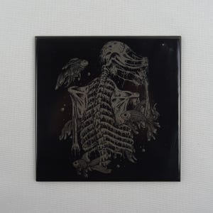 May include: Black and white ceramic tile featuring a detailed illustration of a skeletal figure surrounded by fish. The figure is depicted with a ribcage, spine, and skull. The fish are stylised and have a flowing, organic design.