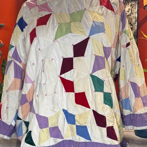 May include: A long-sleeved jacket constructed from a patchwork quilt featuring a white base fabric accented with diamond-shaped patches in red, burgundy, blue, green, yellow, and lavender. The pieces are joined by visible pink cross-stitching.