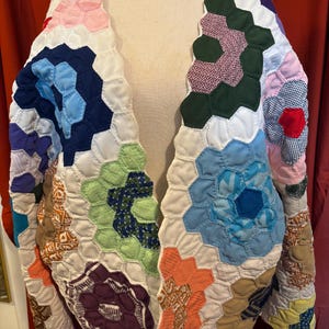 May include: A quilted jacket constructed from numerous multicolored fabric hexagons in shades of blue, green, orange, purple, pink, and white. The garment features a variety of patterns including solids, checks, and prints, all joined together.