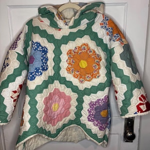 May include: A hooded pullover jacket constructed from a patchwork quilt featuring a hexagonal pattern. The design incorporates various floral and geometric fabric prints in shades of orange, pink, purple, blue, and green, joined by white borders.