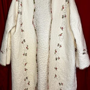May include: A long, cream-colored quilted coat featuring an open front and three-quarter length sleeves. The garment is decorated with embroidered pink rosebuds and green vines along the front edges and sleeve cuffs, finished with a floral trim.
