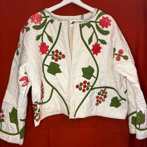 May include: A white quilted jacket featuring an open front and long sleeves. The garment is decorated with an appliqué pattern of green leaves and red circular floral motifs that extend across the front panels and down the sleeves.