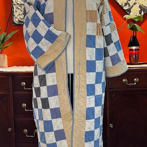 May include: A long, open-front quilted coat featuring a patchwork design of blue, white, and grey checkered squares. The garment has wide sleeves, a solid tan trim along the front opening and hem, and visible stitching throughout the quilted fabric.