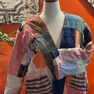 May include: A patchwork jacket displayed on a mannequin. The jacket features a variety of fabrics in pink, blue, orange, and floral patterns. The jacket has a V-neck and long sleeves. The overall style is vintage and handmade.