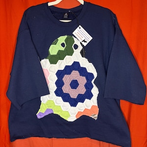May include: A navy blue crewneck sweatshirt featuring a large, central appliqué made of multi-coloured, hexagonal fabric patches in shades of green, white, blue, pink, and orange. A white tag attached to the collar reads HANDMADE BY WOLFGANG.