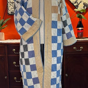 May include: A long, open-front quilted coat featuring a patchwork design of blue, white, and grey checkered squares. The garment has wide sleeves, a solid tan trim along the front opening and hem, and visible stitching throughout the quilted fabric.