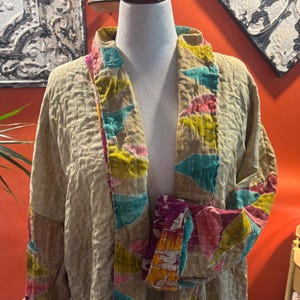 May include: Beige quilted jacket with colorful patchwork trim. The jacket features a relaxed fit and a shawl collar. The trim includes vibrant hues of pink, yellow, teal, and purple. The jacket is displayed on a mannequin.