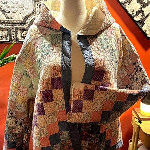 May include: A hooded jacket constructed from a patchwork quilt featuring a variety of floral and geometric patterns in shades of pink, purple, blue, and cream. The garment has long sleeves, a quilted texture, and a solid blue fabric trim.