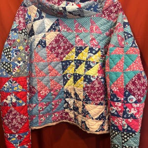 May include: A long-sleeve pullover garment constructed from a patchwork quilt featuring a variety of colorful fabric squares in shades of red, blue, pink, yellow, and teal. The piece has a high, wide collar and a quilted texture throughout.