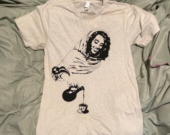 Puede incluir: Camiseta gris claro con un gráfico negro de una persona vertiendo de una botella a una taza de té. La persona lleva un pañuelo en la cabeza y sonríe. La camiseta está hecha de un material suave.