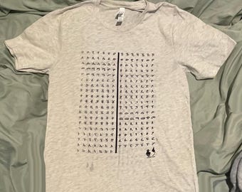 Puede incluir: Una camiseta gris claro con un diseño gráfico negro. El diseño presenta una cuadrícula de símbolos y una línea vertical en el centro. Una pequeña ilustración de una persona está en la parte inferior derecha.