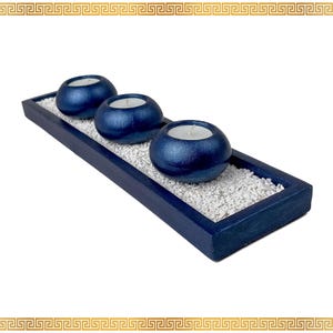 May include: A navy blue wooden candle holder with three spherical, metallic blue candle holders. Each holder contains a white candle. The candle holders sit on a bed of white pebbles within the rectangular tray.