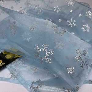 Può includere: Una sciarpa azzurra trasparente con motivi a fiocchi di neve argentati glitterati. La sciarpa è realizzata in un tessuto leggero ed è piegata, mostrando il delicato design. L'etichetta recita "DESO DESIGN".