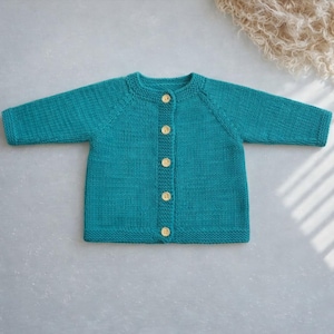 May include: A teal knitted cardigan featuring long sleeves, a round neckline, and a front button closure with five light-coloured wooden buttons. The garment is constructed with a consistent knit texture throughout the body and sleeves.