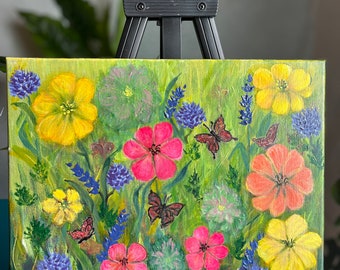 May include: An original painting on canvas featuring a vibrant meadow scene. The artwork showcases colorful flowers in shades of yellow, pink, and purple, with brown butterflies. The painting is displayed on a black easel.