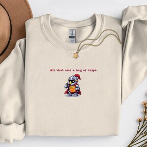 May include: Cream-colored sweatshirt with a crew neck and long sleeves. The front features the text "All that and a bag of chips." below a cartoon knight holding a bag of chips. A gold necklace with a star pendant is draped on the sweatshirt.