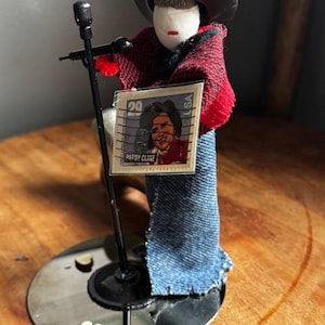 May include: A handcrafted Patsy Cline figurine with a black microphone and a miniature stamp featuring her image. The figure wears a black hat, red and black shawl, and denim clothing. The base is a silver disc with heart-shaped accents.