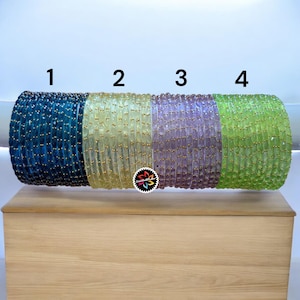 Può includere: Quattro pile di braccialetti di perline blu, gialli, viola e verdi. Ogni pila è adornata da piccole perline color oro. I braccialetti sono esposti su una superficie di legno su sfondo bianco.