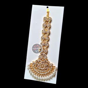 May include: Gold and white jeweled hair accessory with a pearl drop. The accessory is on a white card with the text "Diwali World Fashion".
