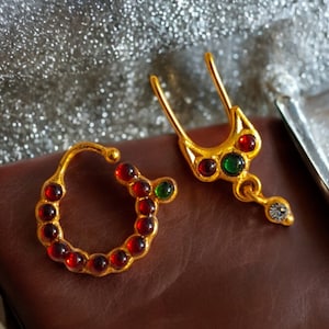 May include: Two gold nose rings with red and green gemstones. One ring is a simple hoop with red gemstones, the other is a more elaborate design with a dangling white gemstone.