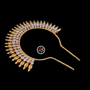 May include: A gold-colored hair accessory with a curved design, adorned with clear crystals and leaf-shaped details. The accessory has two long prongs for securing it in the hair. The background is black.